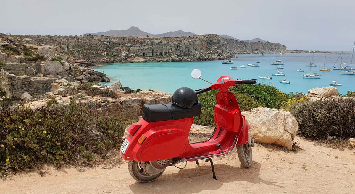 A bright red scooter parked on a dusty clifftop at Cala Rossa, Favignana, with a rocky shoreline and turquoise sea behind. Many small pleasure boats are moored offshore