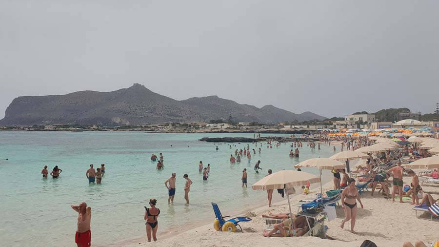 Parasols, sunbeds and beach-goers on the long sandy beach of Lido Burrone on the island of Favignana, with a hilly ridge in the distance