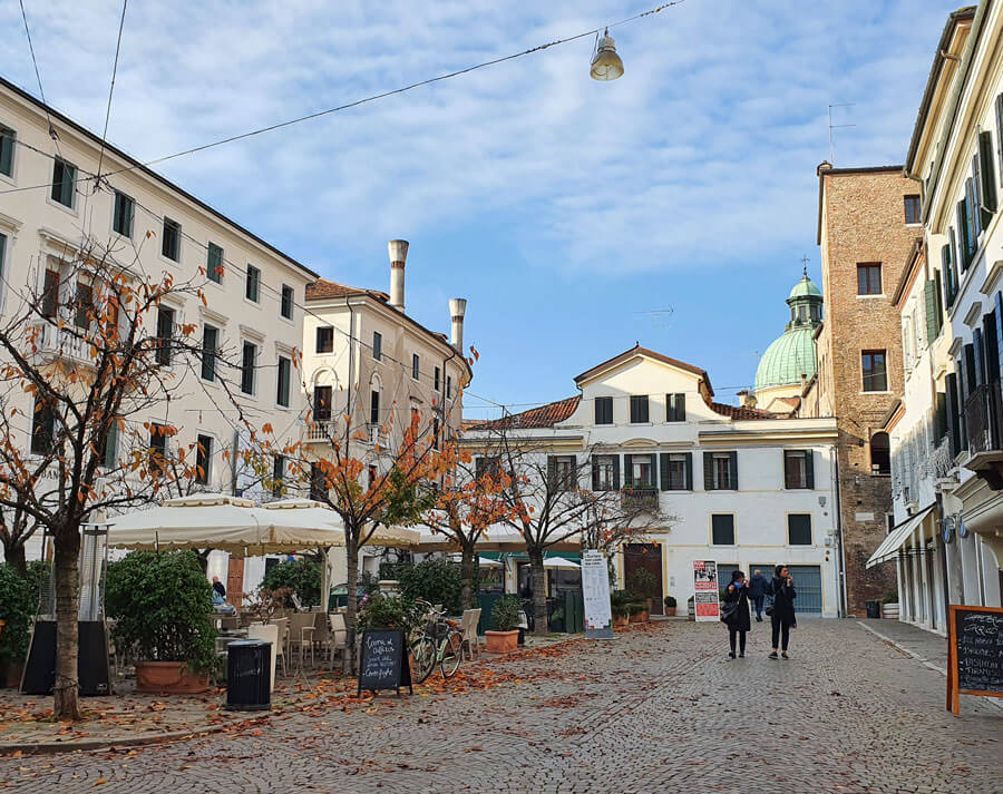 A pretty piazza surrounded by pale buildings with several small trees shedding golden leaves onto the cobbled ground