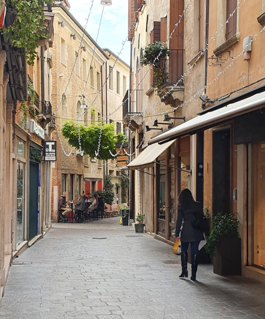 Scenic view of a narrow paved lane with tall buildings, some drinkers outside a bar and a smartly-dressed woman out shopping