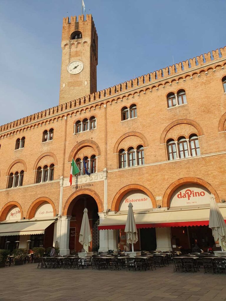 Piazza dei Signori: a tall brick building with merlons and a clocktower, with café tables in front in the piazza