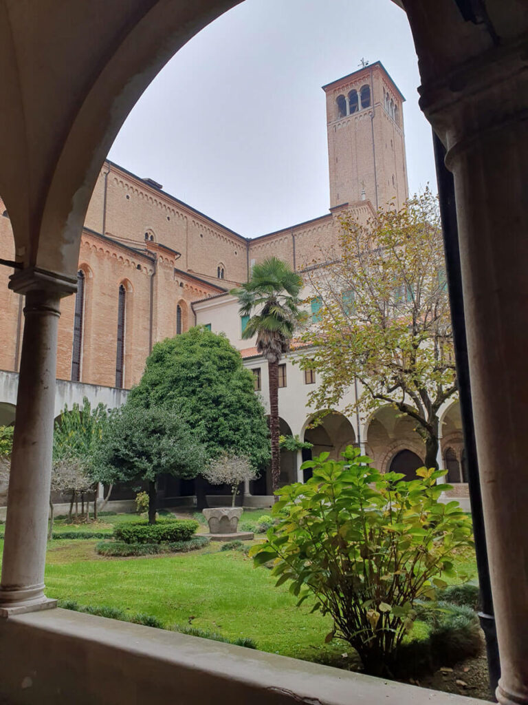 View through an arch supported by narrow columns of a cloister planted with grass, bushes and trees; a church tower in the background